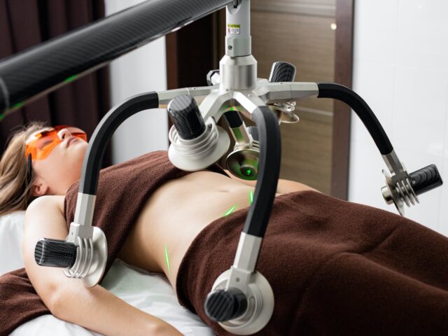 Photo by Dmitriy Ganin Female client lying on table under lasers of modern weight loss machine on belly during liposuction procedure in medical clinic